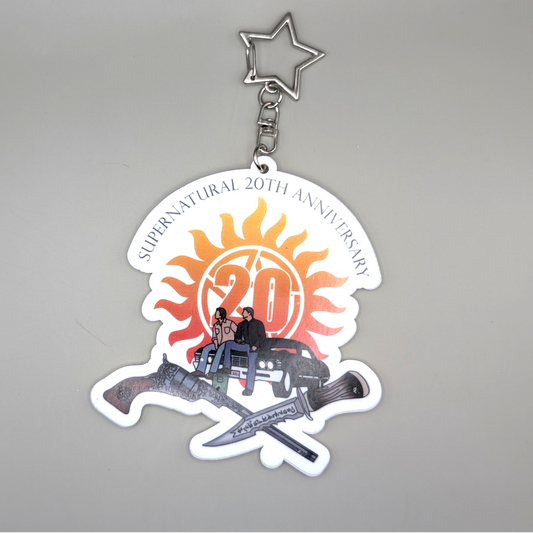White Keychain with colorful logo for the 20th Anniversary of Supernatural. Sam and Dean sitting on Baby and the Cold and Ruby's Blade beneath it.